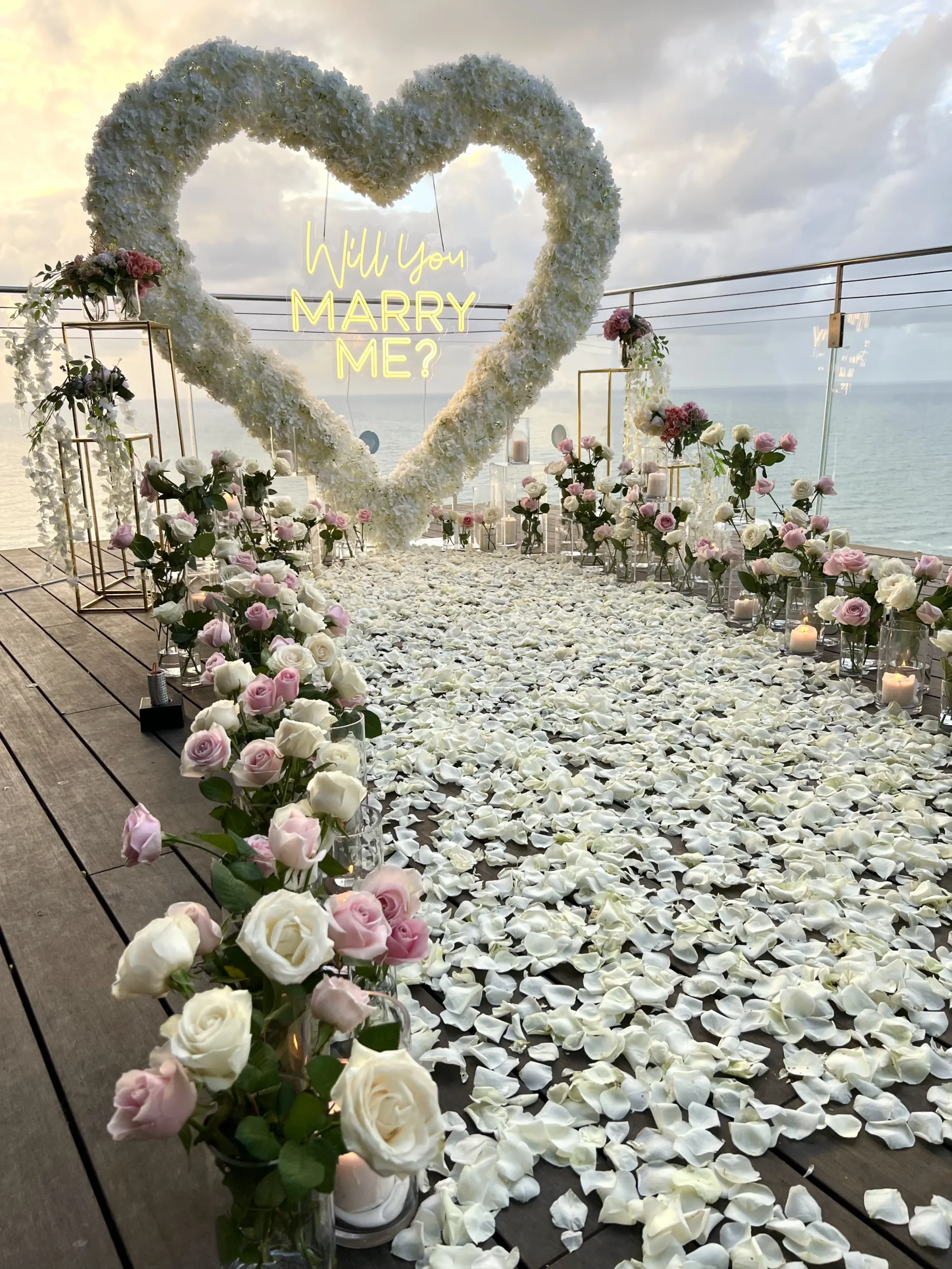 הצעת נישואין מהחלומות - שביל עוצמתי של פרחים אמיתיים בגוונים לבן וורוד , מוביל ללב ענק מרחף ושלט מואר בכיתוב "'will you marry me?" מול שקיעה עוצרת נשימה. עיצוב מרגש, רומנטי ומדויק עד הפרח האחרון.