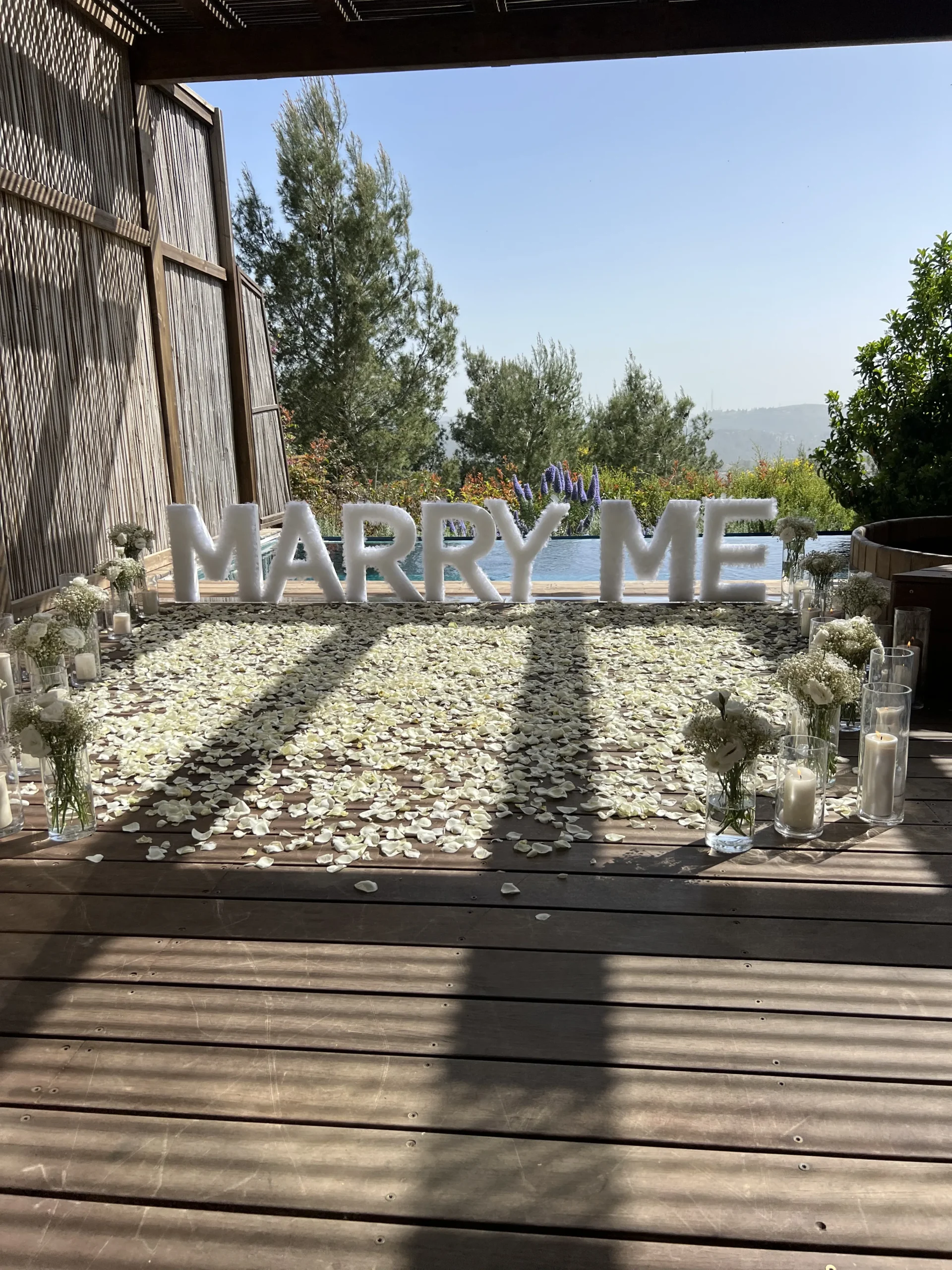 הצעת נישואין בעיצוב אותיות MARRY ME , זרי פרחים חיים , נרות ועלי כותרת המשדרגים את החלל.