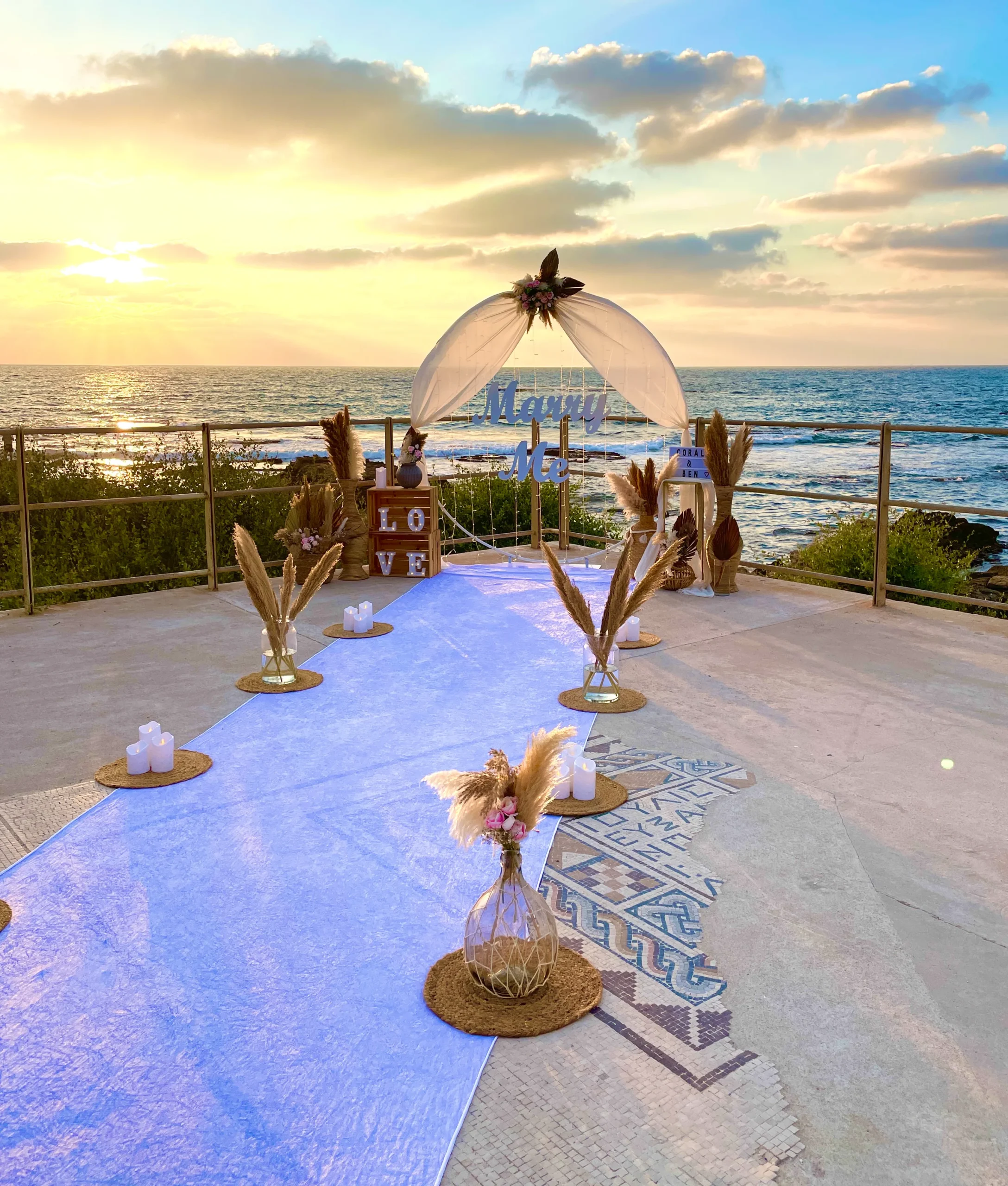 עיצוב להצעת נישואין בסגנון קלאסי ונקי , עם נגיעות בוהו - המשלבת שביל הליכה מואר בנרות ופרחים המוביל לעמדה עוצמתית מעוטרת בתאורה רכה, בדים תואמים וזרי פרחים. שלט לבן בכיתוב " marry me". שילוב של אלגנטיות, רגש ונוכחות בלתי נשכחת !