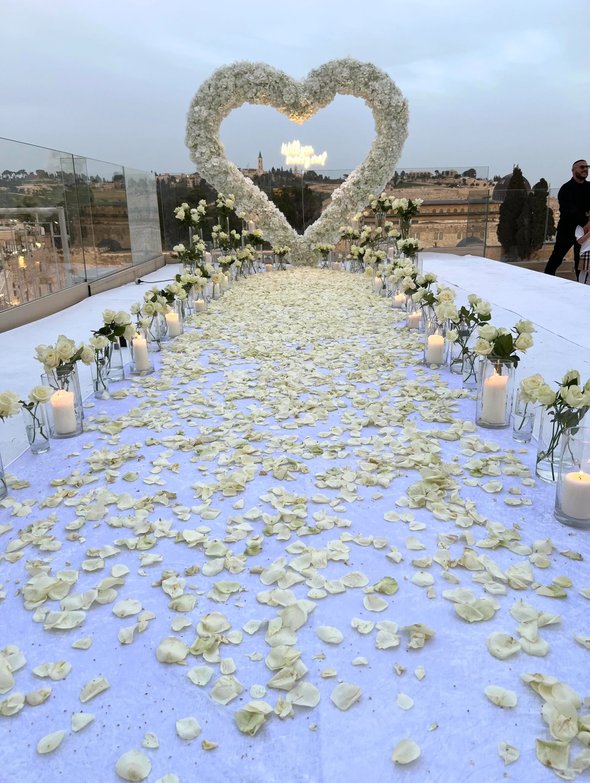 הצעת נישואין מהחלומות - שביל עוצמתי של פרחים אמיתיים בגוון שמנת, מוביל ללב ענק מרחף ושלט מואר בכיתוב "'will you marry me?" בלב הכותל המערבי. עיצוב מרגש, רומנטי ומדויק עד הפרח האחרון.