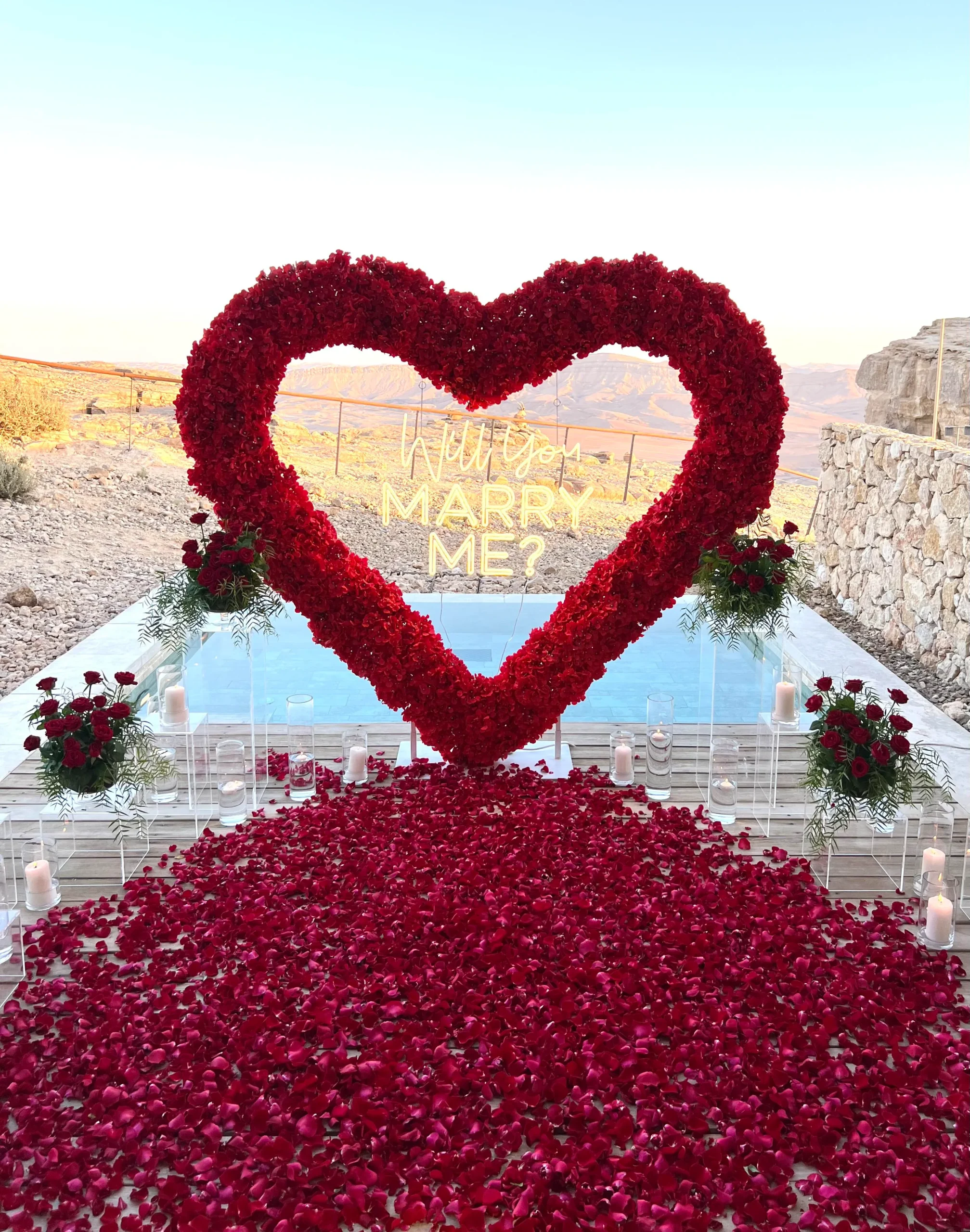 הצעת נישואין בעיצוב דרמטי ורומנטי במיוחד – לב ענק וייחודי מפרחים אדומים, סידור וורדים טריים כעלי כותרת, ריבוי וואזות ונרות , ושזירת ארבעה זרי פרחים חיים משני צדי הלב. עיצוב שמדבר אהבה – באדום עוצמתי ובדיוק מושלם.