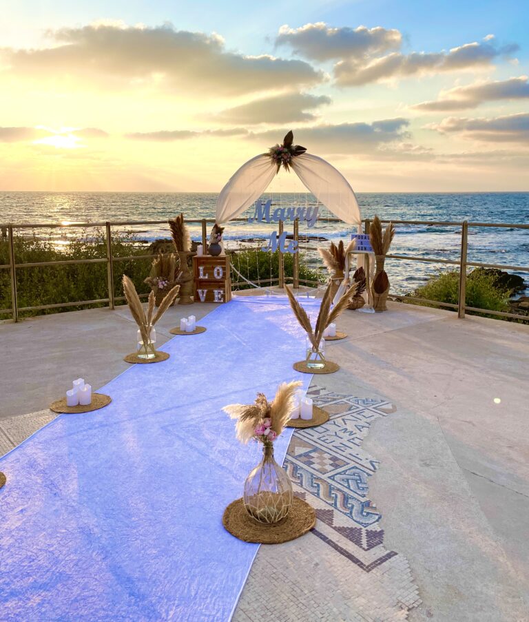 עיצוב להצעת נישואין בסגנון קלאסי ונקי , עם נגיעות בוהו - המשלבת שביל הליכה מואר בנרות ופרחים המוביל לעמדה עוצמתית מעוטרת בתאורה רכה, בדים תואמים וזרי פרחים. שלט לבן בכיתוב " marry me". שילוב של אלגנטיות, רגש ונוכחות בלתי נשכחת !