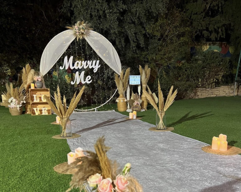 עיצוב להצעת נישואין בסגנון קלאסי ונקי , עם נגיעות בוהו - המשלבת שביל הליכה מואר בנרות ופרחים המוביל לעמדה עוצמתית מעוטרת בתאורה רכה, בדים תואמים וזרי פרחים. שלט לבן בכיתוב " marry me". שילוב של אלגנטיות, רגש ונוכחות בלתי נשכחת !